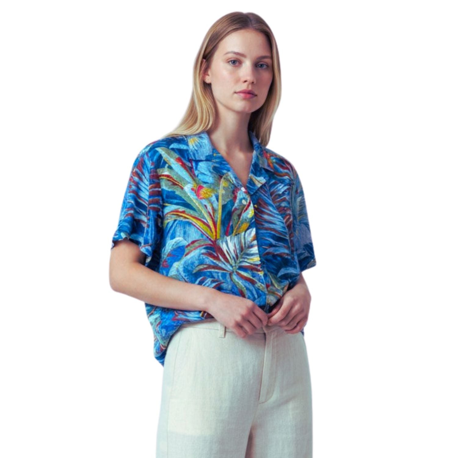 A woman modeling a short-sleeve tropical print blouse with blue, green, yellow, and red palm leaves, paired with light beige linen trousers, against a solid white background.
