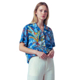 A woman modeling a short-sleeve tropical print blouse with blue, green, yellow, and red palm leaves, paired with light beige linen trousers, against a solid white background.