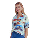 A woman modeling a short-sleeve button-up shirt with a colorful retro racing and travel-themed print on a light background, paired with blue jeans against a plain white studio background.