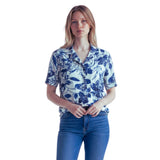 A woman modeling a short-sleeve button-up shirt with a blue floral print on a white background, paired with blue jeans, against a plain white studio background.