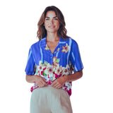 A woman modeling a short-sleeve button-up shirt with a vibrant blue base, vertical white stripes, and a colorful tropical floral print at the hem and collar, paired with light beige trousers against a solid white background.
