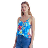 A woman modeling a bright blue floral spaghetti-strap camisole top with red, yellow, and white flowers, paired with blue jeans, against a plain white background.