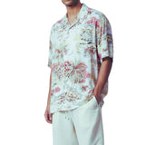 A man wearing a short-sleeve button-up shirt with a tropical floral and palm tree print in pink, green, and beige tones, paired with off-white drawstring pants, against a plain white background.