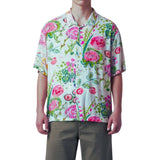 A man wearing a short-sleeve button-up shirt with a vibrant pink, green, and blue floral watercolor print on a white base, paired with olive green pants, against a plain white background.