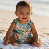A baby sitting on a white towel on a sandy beach wearing a sleeveless light blue floral swimsuit.