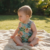 A baby sitting outdoors on a cream knit blanket, wearing a sleeveless floral romper with a light blue background and orange, pink, and green tropical flowers.