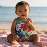 A baby sitting on a pink blanket on a sandy beach, wearing a colorful tropical print sleeveless swimsuit.