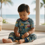 A toddler sitting on a light carpet indoors near large windows, wearing a short-sleeve pajama set with a colorful ocean-themed print in blue, teal, yellow, pink, and beige. The child is holding wooden blocks.
