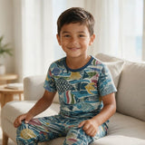 A young boy smiling while sitting on a light beige sofa, wearing a short-sleeve pajama set with a colorful fish pattern in blue, yellow, and red.