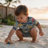 A toddler crouches on a sandy beach wearing a short-sleeve swim shirt and matching swim trunks with a colorful fish pattern. The child is interacting with a small toy in the sand, with a wooden fence and palm tree blurred in the background.