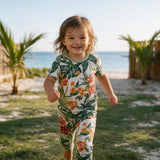 A young child wearing a short-sleeve tropical floral print one-piece jumpsuit, smiling and running outdoors on a grassy lawn with palm trees and the ocean in the background.