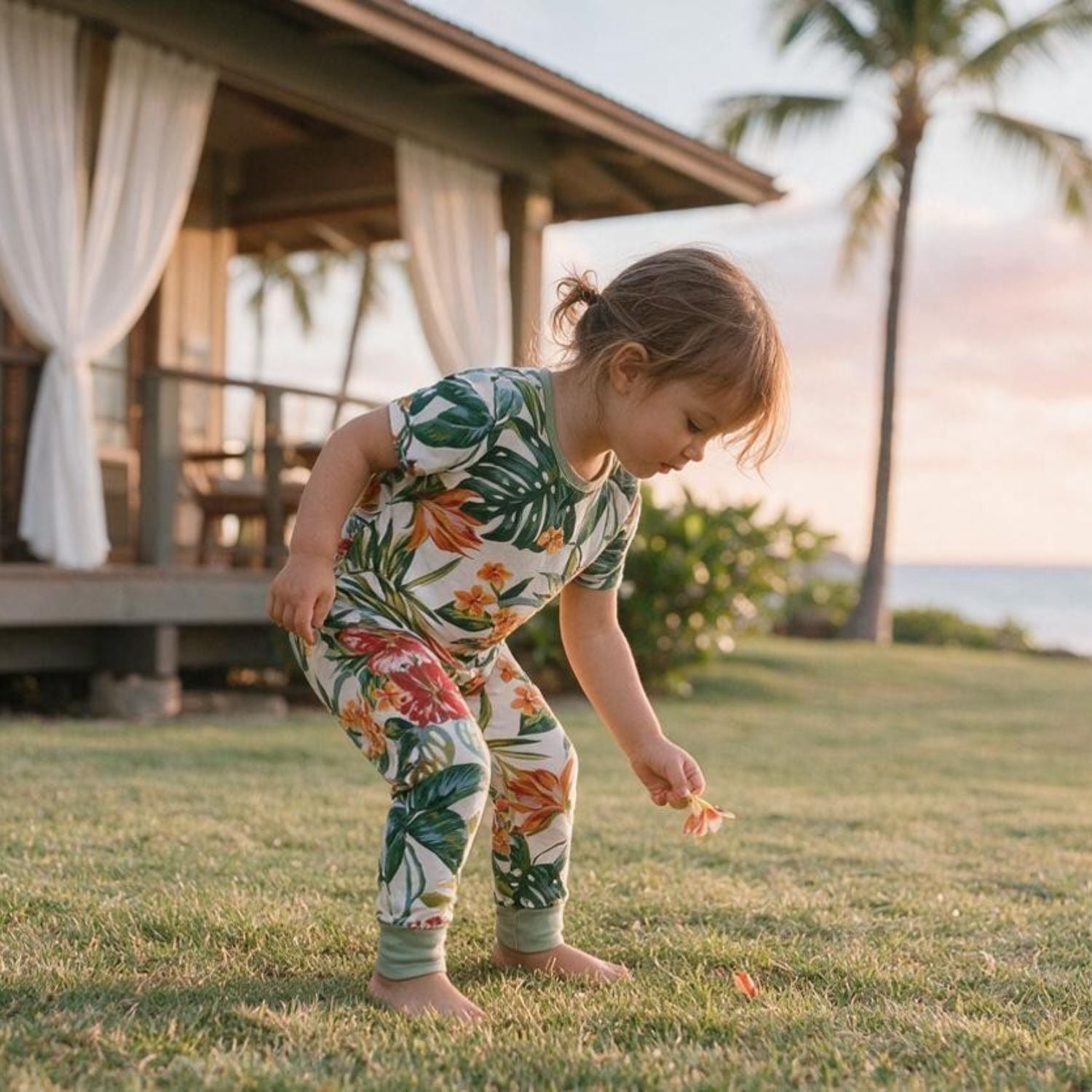 A toddler wearing a short-sleeve tropical-print one-piece jumpsuit bends down on a grassy lawn near a house and palm trees.