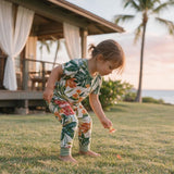 A toddler wearing a short-sleeve tropical-print one-piece jumpsuit bends down on a grassy lawn near a house and palm trees.