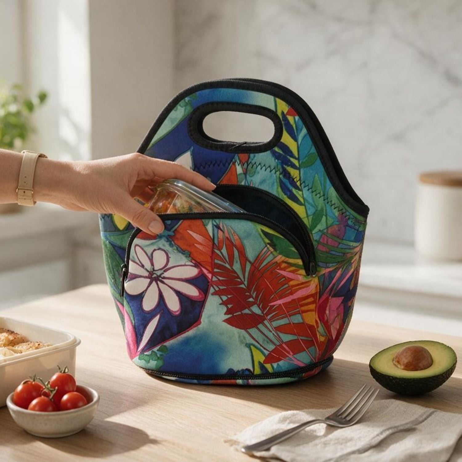 A colorful insulated lunch bag with a tropical floral and leaf print sits on a wooden table. A hand is placing a glass container into the bag's front pocket.