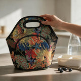 A hand holds the top handle of a black insulated lunch bag with a colorful coral and starfish print, set on a kitchen counter next to a lunchbox and blueberries.