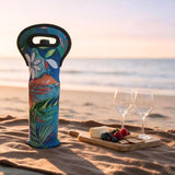 A colorful tropical floral insulated bottle with a black handle sits on a sandy beach next to a wooden charcuterie board with cheese, crackers, and two wine glasses, with the ocean and sunset in the background.