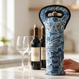 A blue and white patterned wine bottle carrier with a black handle is shown on a kitchen counter next to wine bottles and wine glasses, with a hand holding the carrier.