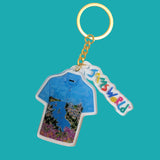 A Jaws World keychain featuring a molded acrylic keyring shaped like a blue Hawaiian shirt with a colorful coastal landscape illustration, attached to a gold-tone chain and a dangling tag that reads 'JAWS WORLD' in multicolored letters, set against a solid teal background.