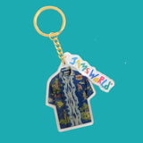 A keychain featuring a die-cut acrylic charm shaped like a blue Hawaiian shirt with a tropical floral print and waterfall graphic, attached to a gold-tone split ring and chain. A colorful script tag on the charm reads 'Jinsworld'.