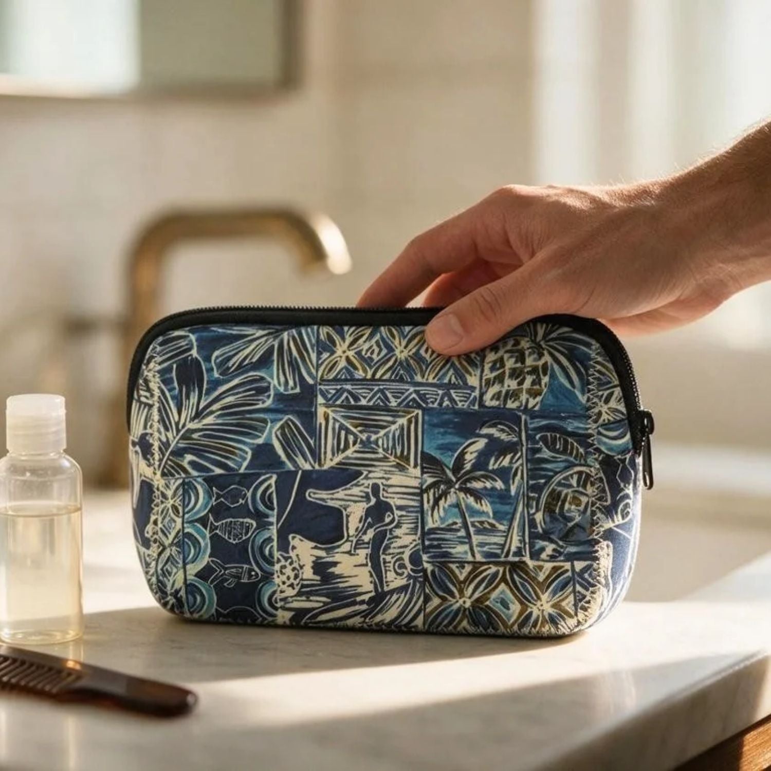 A hand holds a zippered toiletry bag with a blue and cream tropical print, set on a bathroom counter next to a small bottle and comb.