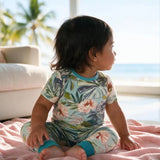 A toddler sitting on a pink blanket in front of a window, wearing a short-sleeve matching pajama set with a tropical floral print and teal cuffs.