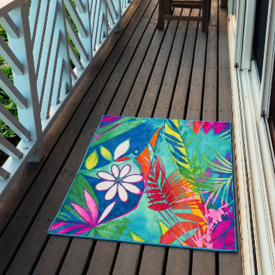 A rectangular outdoor area rug with a vibrant tropical floral and leaf pattern in bright teal, pink, orange, green, and purple, displayed on a dark wooden deck next to a white railing.