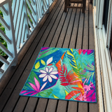 A rectangular outdoor area rug with a vibrant tropical floral and leaf pattern in bright teal, pink, orange, green, and purple, displayed on a dark wooden deck next to a white railing.