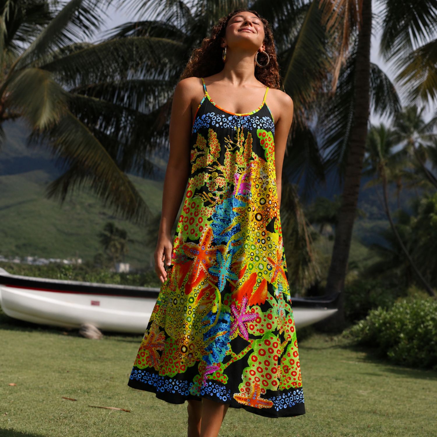 A woman wearing a vibrant, multicolored spaghetti-strap midi dress with a tropical floral print, standing outdoors on a grassy lawn with palm trees and a boat in the background.