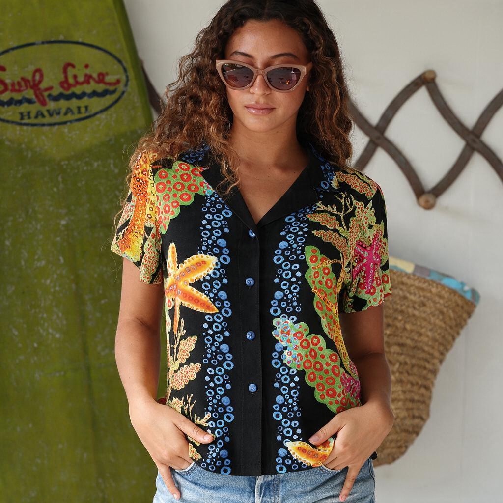 A woman wearing a short-sleeve button-up shirt with a black base and a colorful tropical floral pattern, standing in front of a surfboard and a woven basket.