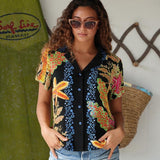 A woman wearing a short-sleeve button-up shirt with a black base and a colorful tropical floral pattern, standing in front of a surfboard and a woven basket.