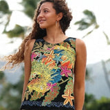 A woman wearing a sleeveless black tank top with a vibrant, multicolored tropical print featuring starfish, coral, and green leaves, set against a blurred outdoor background of palm trees and mountains.