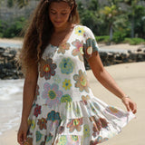 Una mujer con un vestido blanco de volantes y estampado floral colorido se encuentra en una playa de arena, mostrando la caída de la falda. El vestido tiene mangas cortas, escote redondo y un vibrante estampado floral multicolor.