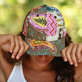A woman outdoors wearing a colorful patchwork baseball cap with bohemian patterns, holding the brim down over her eyes.
