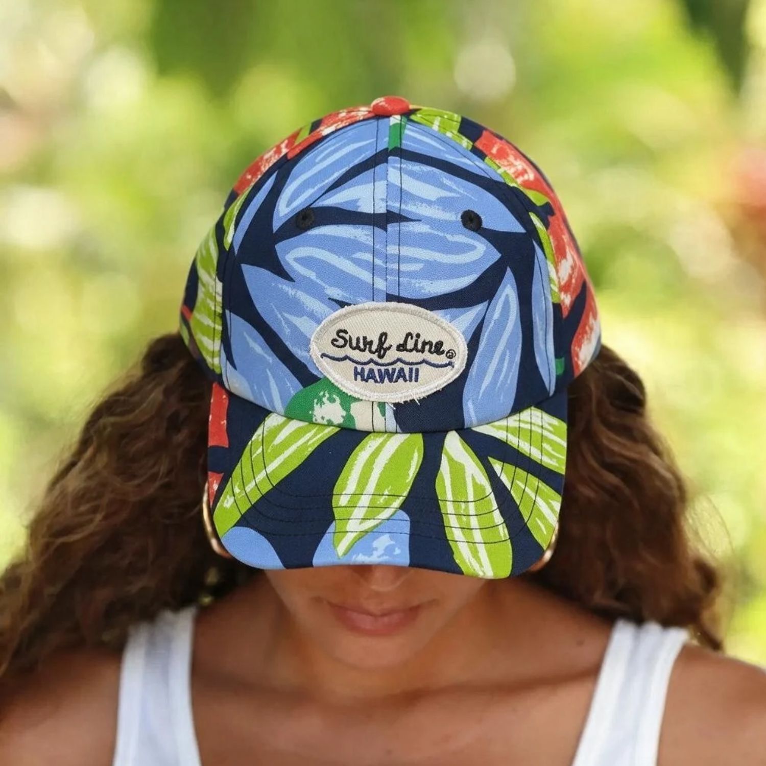 A woman wearing a colorful Surf Line Hawaii baseball cap with a blue and green tropical leaf pattern, photographed outdoors with a blurred green background.