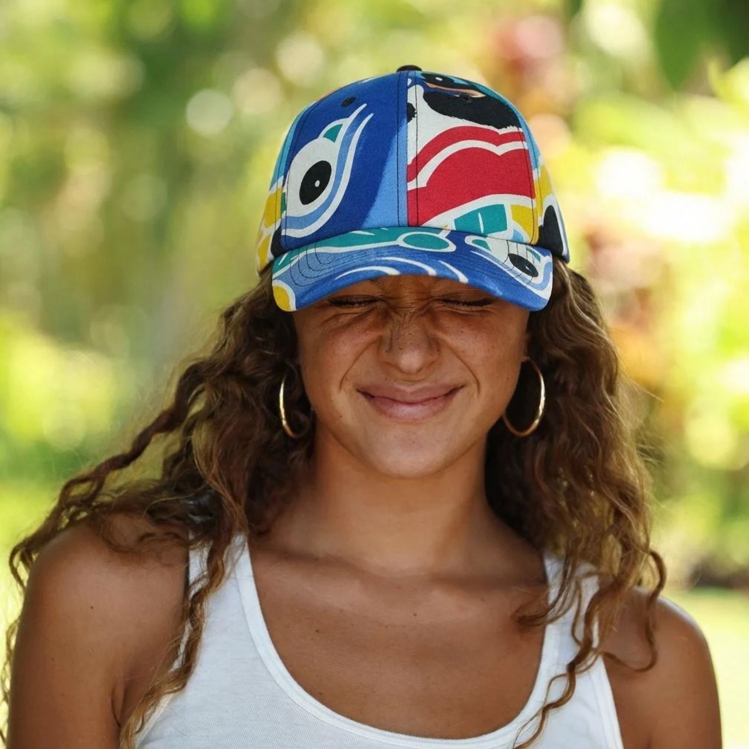 A woman outdoors wearing a colorful patterned baseball cap with blue, red, yellow, and white abstract designs, along with gold hoop earrings and a white tank top.