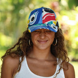 A woman outdoors wearing a colorful patterned baseball cap with blue, red, yellow, and white abstract designs, along with gold hoop earrings and a white tank top.