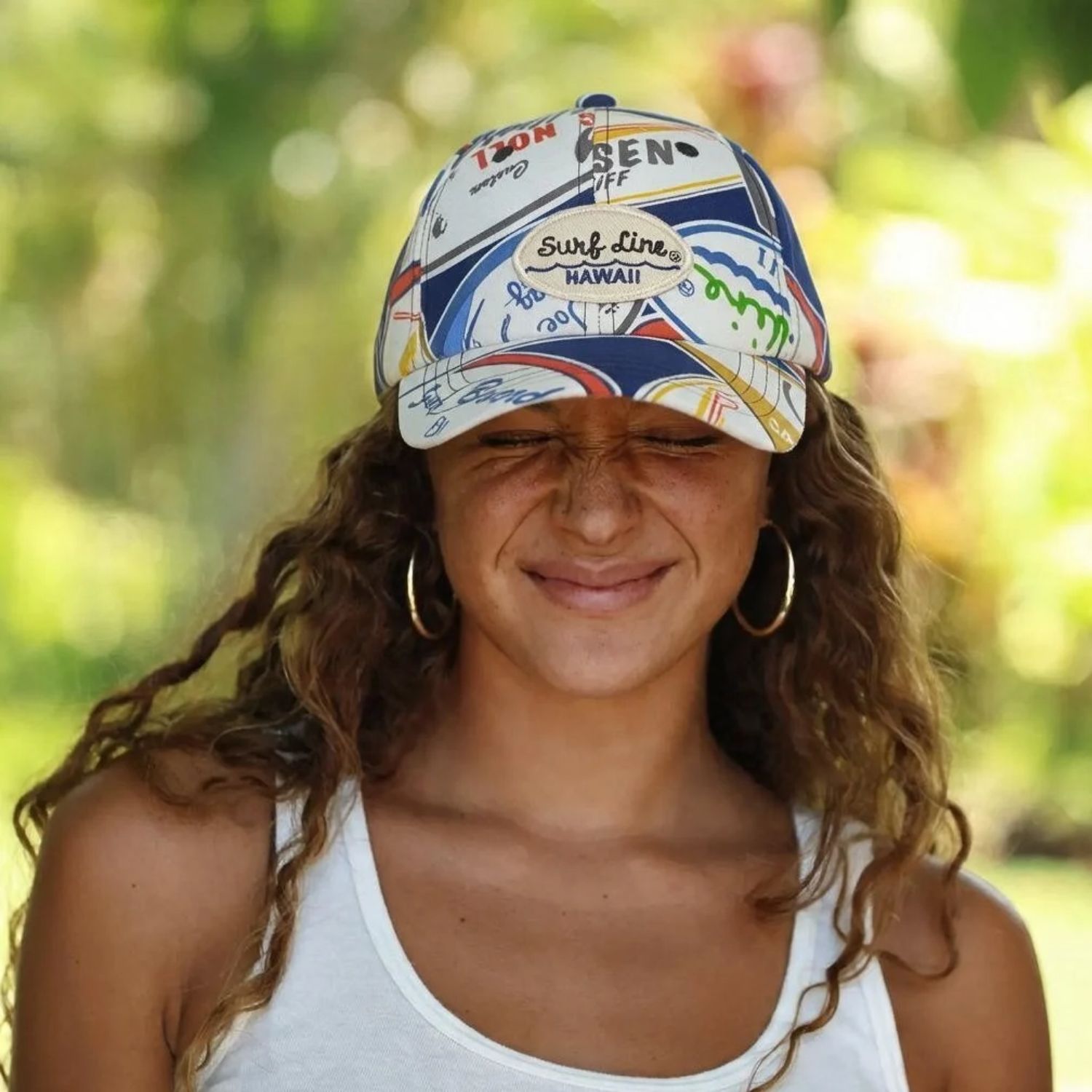 A woman outdoors wearing a white baseball cap covered in colorful 'Sun Line Hawaii' and other travel-style graphics, along with gold hoop earrings and a white tank top, against a blurred green background.