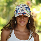 A woman outdoors wearing a white baseball cap covered in colorful 'Sun Line Hawaii' and other travel-style graphics, along with gold hoop earrings and a white tank top, against a blurred green background.