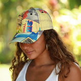 A woman wearing a baseball cap with a vintage-style collage print featuring blue, red, yellow, and black elements, set against a blurred green outdoor background.