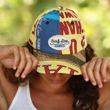 A woman wearing a multicolor Surf Line Hawaii baseball cap, holding the brim down over her eyes. The cap features a split design with a blue and yellow graphic top and a camouflage-patterned brim.