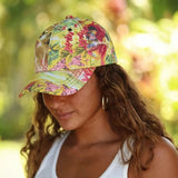 A woman wearing a vibrant tropical floral baseball cap with a curved brim, photographed outdoors against a soft, blurred green and yellow background.