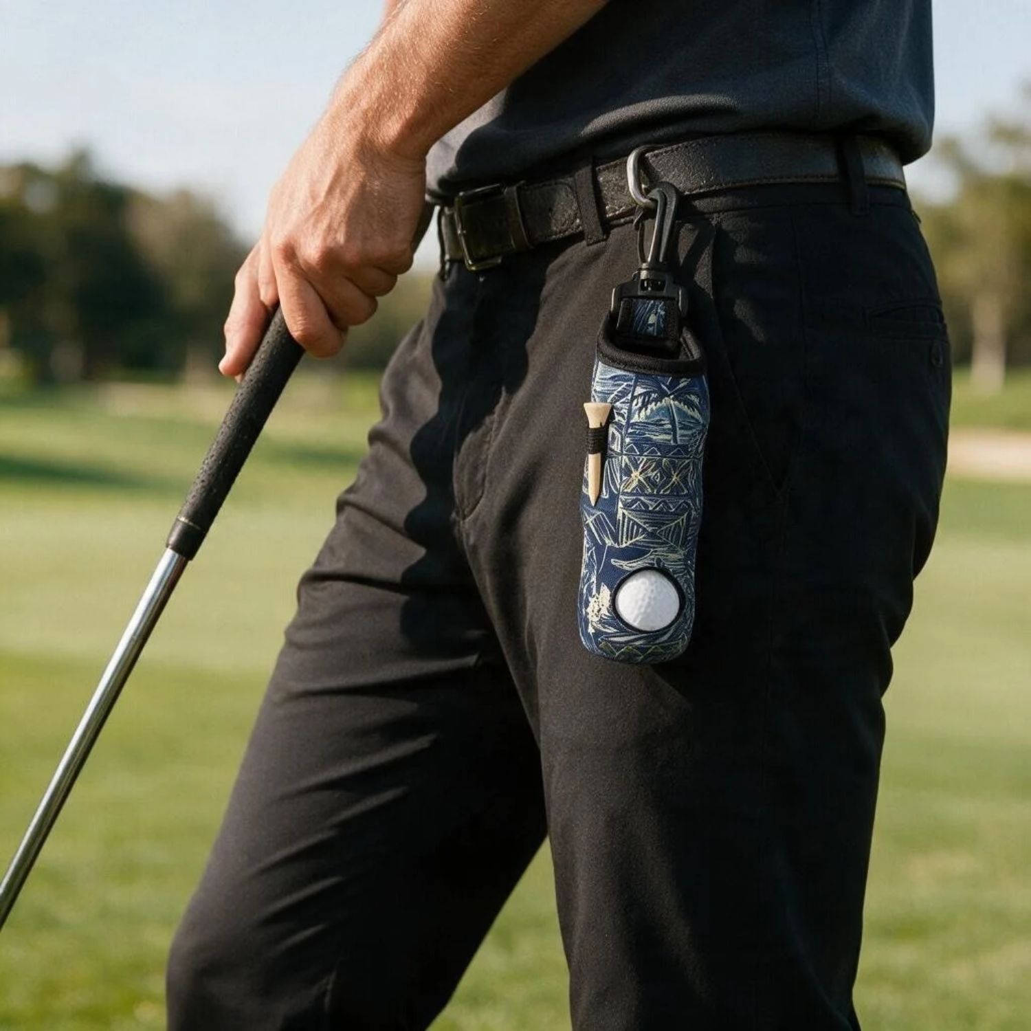 A close-up of a person on a golf course wearing black pants and a dark shirt, holding a golf club. A patterned blue and white golf accessory pouch with a black clip is attached to the belt loop.