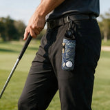 A close-up of a person on a golf course wearing black pants and a dark shirt, holding a golf club. A patterned blue and white golf accessory pouch with a black clip is attached to the belt loop.