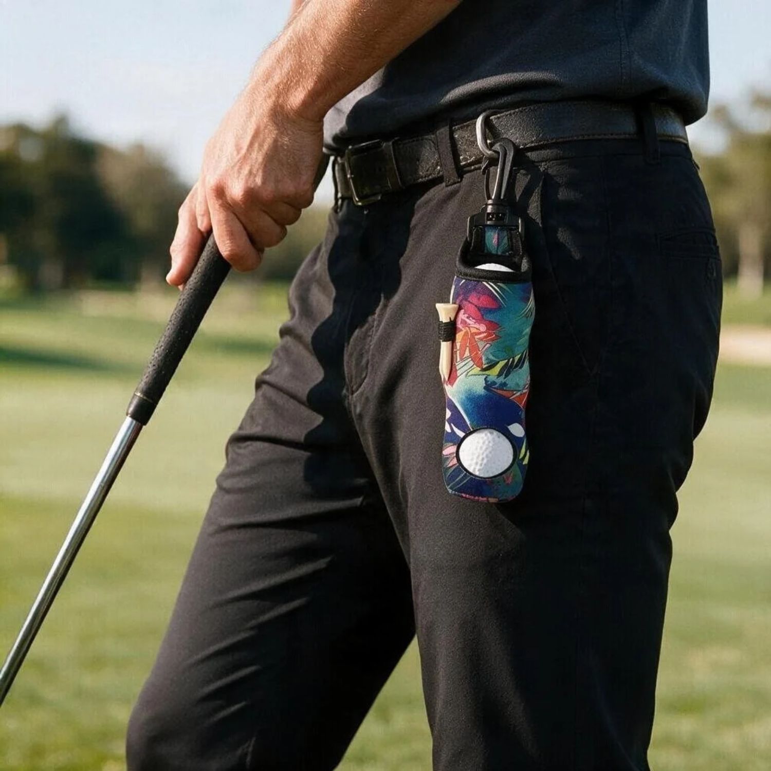 A close-up of a person on a golf course wearing black pants and a dark shirt, holding a golf club. A colorful, patterned golf accessory with a white golf ball attached to a black clip hangs from the belt on the thigh.