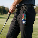 A close-up of a person on a golf course wearing black pants and a dark shirt, holding a golf club. A colorful, patterned golf accessory with a white golf ball attached to a black clip hangs from the belt on the thigh.