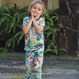A young girl wearing a matching short-sleeve t-shirt and long-sleeve pants pajama set with a vibrant tropical floral print, posing outdoors in front of a stone wall and green plants.
