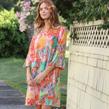 A woman wearing a colorful tropical leaf print kaftan dress stands outdoors next to a light-colored wooden fence with green foliage and pink flowers in the background.