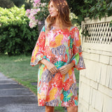 A woman wearing a vibrant, multicolored tropical print kaftan dress with wide flared sleeves, standing outdoors next to a white wooden lattice fence and green bushes.
