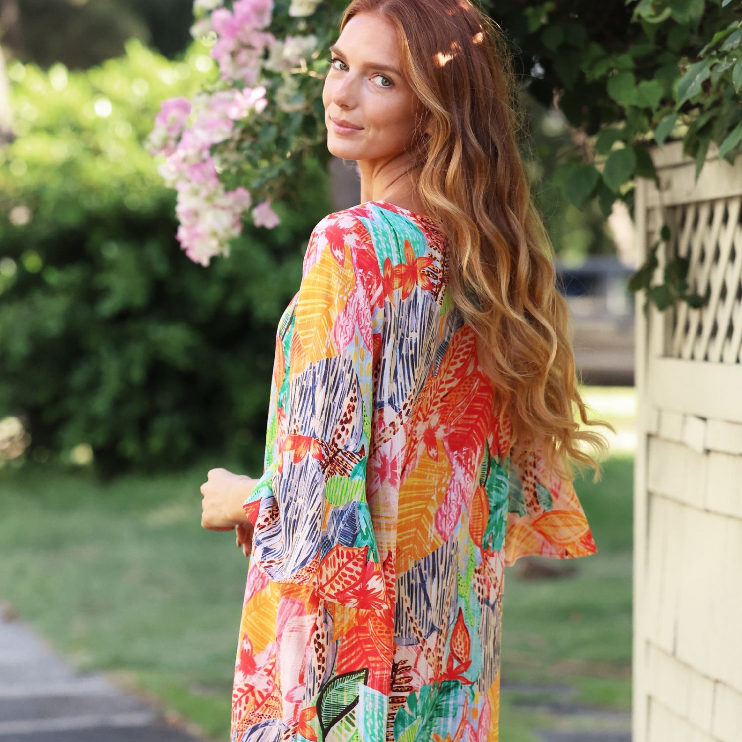 A woman wearing a vibrant, multicolored tropical leaf print kaftan dress, photographed outdoors in a garden setting.