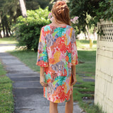 A woman viewed from behind wearing a colorful, short-sleeved tiered dress with a vibrant tropical floral and leaf print, standing on a stone path in a garden setting.